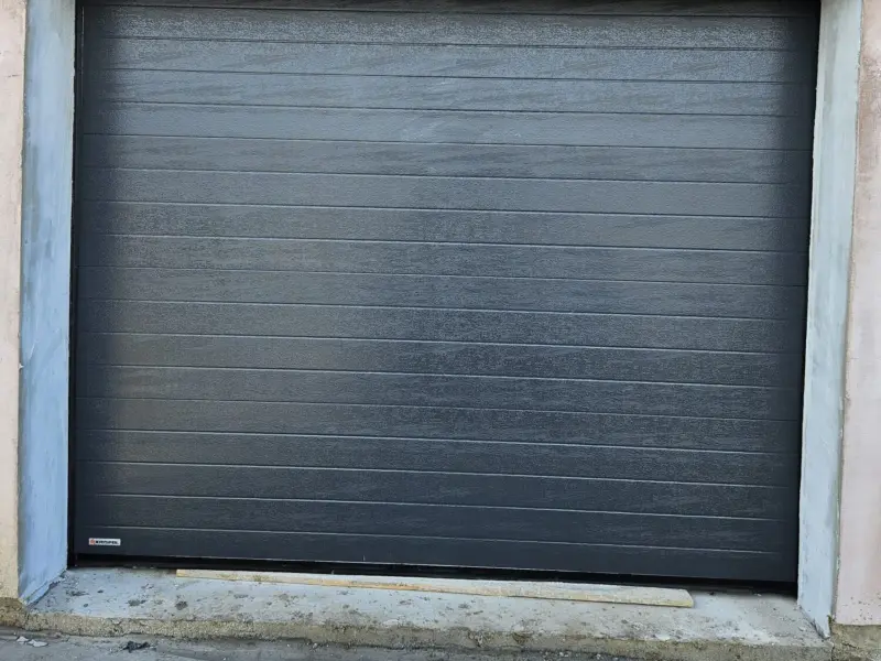 Ușă de garaj secțională KRISPOL antracit, cu lamelă îngustă și structură Woodgrain, montată pe fațadă